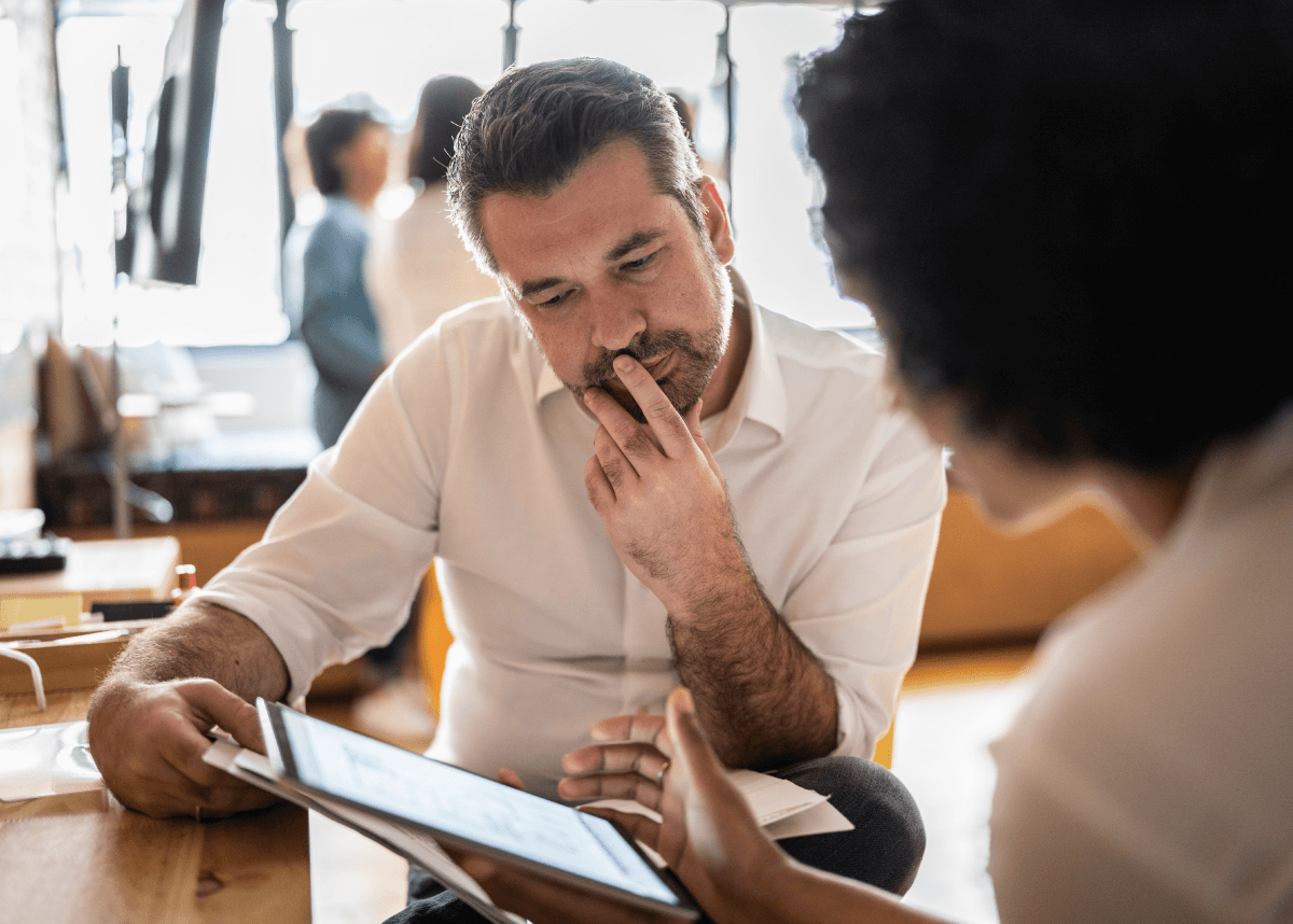 A man with a thoughtful expression looking at a tablet, sitting at a table indoors with blurred people in the background.